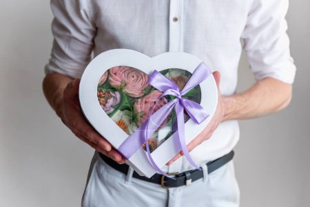 Photo for Faceless man holds box shape of heart of marshmallow sweet flowers in hands. Gift with purple ribbon. Delicate sugarless roses on light background. Small business at home. Hobby baking. Holiday gift - Royalty Free Image