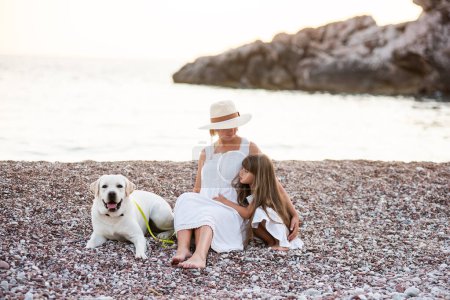 Photo for Pregnant mother with little daughter are sitting along the sand of the beach near the sea with golden retriever. Millennial woman holds dog on leash, girl by the hand. Concept of traveling with pets - Royalty Free Image
