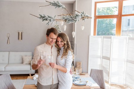 Close-up portrait of loving, young fashion couple with glasses of champagne in Christmas. Trendy interior in Scandinavian style with panoramic window and snow-covered branch. Lovers hugging, tactility