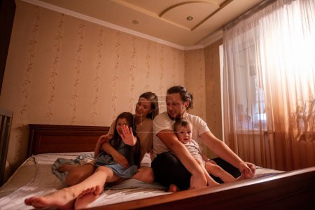 Photo for Portrait of diverse family in bedroom on bed in morning sun. Eldest daughter protests, putting her hand forward with Stop sign. Father holds baby son in arms, mother looks at girl. Lifestyle cozy - Royalty Free Image