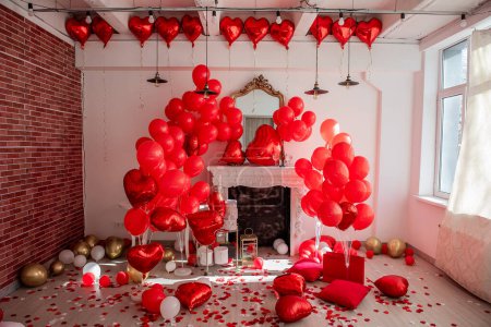 Téléchargez les photos : Décoration de chambre chic pour la Saint-Valentin. Cheminée près du mur blanc décorée de ballons rouges en forme de coeur, sur le sol cadeaux dans des boîtes de vacances, sacs, confettis, pétales de rose dispersés. Vin avec verres - en image libre de droit