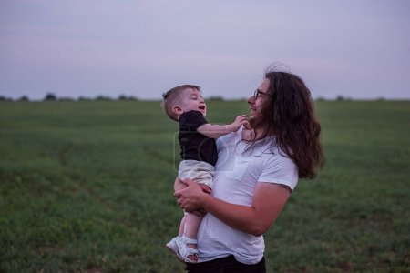 Close up portrait of young father with long hair, beard, glasses holds little son, who playfully raises his arm, both enjoying casual moment of vast green field. Beaming toddler boy with style man