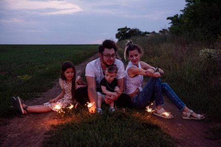 Familia de la diversidad de cuatro jugadas con bengalas en el camino rural, compartiendo un momento acogedor mientras crepúsculo se instala. Brillante celebración en la naturaleza