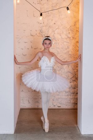 Poised young ballerina stands en pointe in hallway arch with warm garlands of lamps, elegant costume and focused demeanor embodying the spirit of classical ballet.
