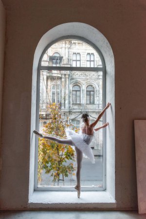 Ballerina poses en pointe, white tutu within an arched window frame, juxtaposing the refined beauty of ballet with the urban landscape outside. Urban Ballet Elegance