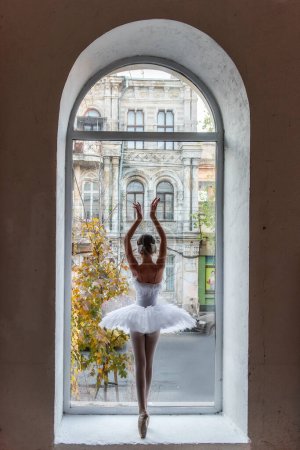 Bailarina posa en punta, tutú blanco dentro de un marco de ventana arqueada, yuxtaponiendo la refinada belleza del ballet con el paisaje urbano exterior. Ballet urbano Elegancia