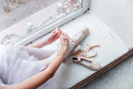 Vista aérea captura a una bailarina sentada junto a una ventana brillante, atando meticulosamente sus zapatos puntiagudos en preparación para la danza, rodeada por el paisaje urbano exterior.