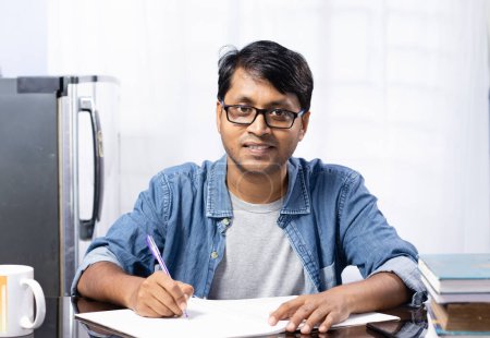 Ein junger indischer Mann blickt mit lächelndem Gesicht in die Kamera, während er zu Hause auf weißem Hintergrund studiert