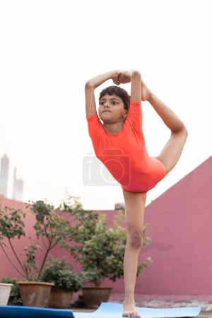 Téléchargez les photos : Une fille indienne enfant pratiquant le yoga dandayamana Purna Dhanurasana sur tapis de yoga en plein air - en image libre de droit