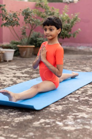 Téléchargez les photos : Une fille indienne enfant pratiquant hanumanasana yoga sur tapis de yoga en plein air - en image libre de droit