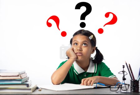 Ein hübsches indisches Mädchen am Lerntisch mit verwirrtem Gesicht, das Fragezeichen auf weißem Hintergrund betrachtet