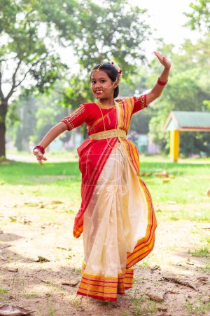 Photo for A pretty beautiful Indian girl child in traditional dancing dress, red saree and jewellery posing dance steps outdoors with smiling face - Royalty Free Image