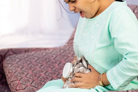 Una mujer india que aplica bolsa de agua caliente en su vientre para aliviar el dolor sentada en un sofá
