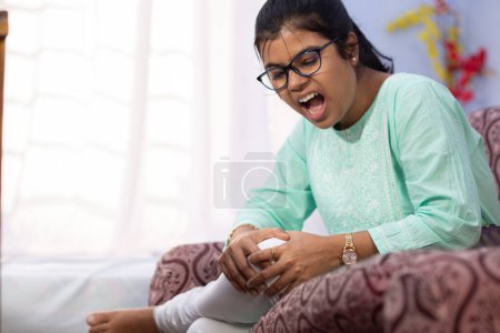 An Indian woman holding her knee in pain showing painful expression sitting on sofa