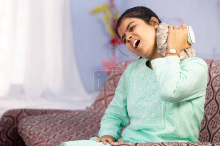 An Indian woman using hot water bag on her neck for pain relief showing painful expression sitting on couch indoors