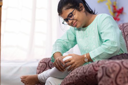 An Indian woman holding her knee in pain showing painful expression sitting on sofa