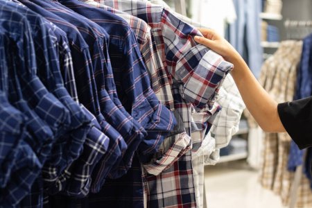 Selektiver Fokus auf Kleidungsstücke, die an Kleiderbügeln hängen und menschliche Hand in einem Einkaufszentrum