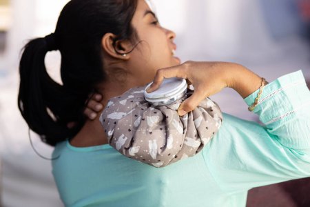 An Indian woman using hot water bag for shoulder pain relief on white background