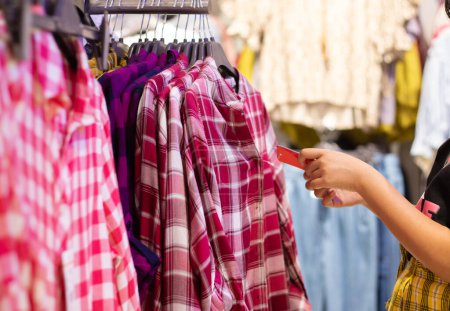 Selektiver Fokus auf Kleidungsstücke, die an Kleiderbügeln hängen und menschliche Hand, die Preisschilder in einem Einkaufszentrum überprüft