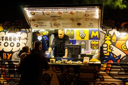 Foto de Barcelona, España-25 de noviembre de 2023. Grupo de personas que se divierten mientras comen delante de un camión modificado para el servicio de comida rápida móvil. Camión de comida - Imagen libre de derechos