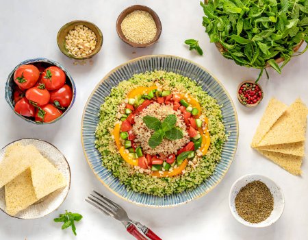 A vibrant bowl of fresh tabbouleh salad garnished with a sprig of mint, surrounded by ingredients like tomatoes and olive oil, set on a white surface.