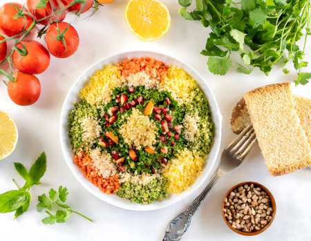 Eine lebhafte Schüssel mit frischem Tabbouleh-Salat, garniert mit einem Zweig Minze, umgeben von Zutaten wie Tomaten und Olivenöl, auf einer weißen Oberfläche.