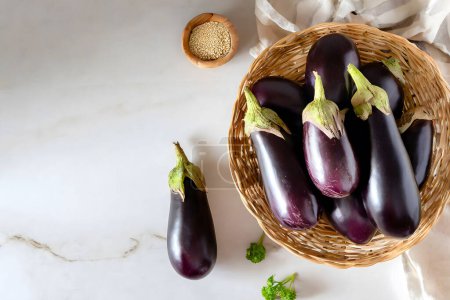 Top view of vibrant purple eggplants in a rustic woven basket beside a bowl of seeds on a marble background, evoking a farm-to-table concept