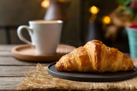 A delicious buttery croissant served on a black plate with a cup of coffee in the background on a wooden surface