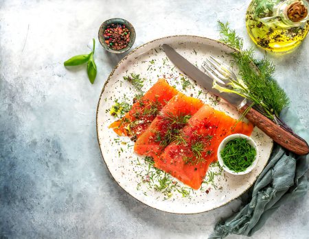 Top view of a plate of smoked salmon adorned with fresh dill, spices, and a drizzle of olive oil on a textured background. Gravlax