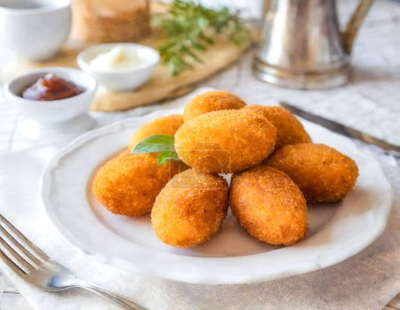 A plate of fried croquettes sits on a table with a fork and knife. The table is set with a white plate