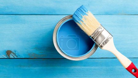 A blue paint can sits on a wooden surface with a paintbrush next to it. The blue paint is ready to be used for painting