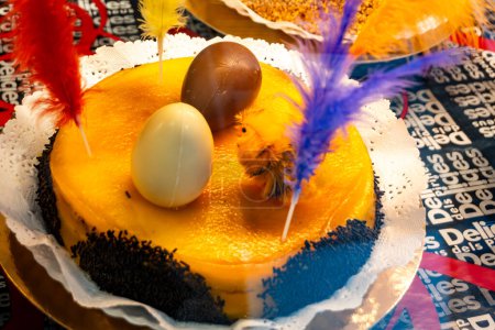 a mona de pascua, a cake eaten in Spain on Easter Monday, topped with a chocolate chick, on a rustic wooden surface full of decorated eggs and feathers of different colors
