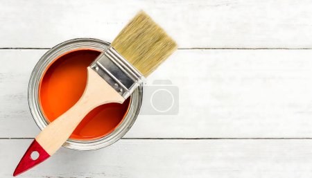 Orange paint can with a paintbrush on top of it on a white wooden table. Close-up