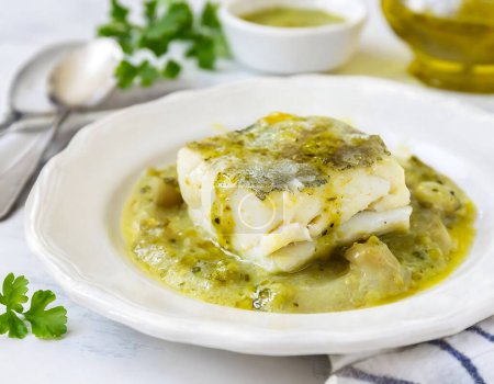 Plate of hake in green sauce served on a white plate elegantly placed on a wooden table.