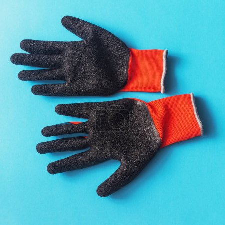 Photo for Overhead view of a pair of knitted red gloves with black latex covered palms facing up against a blue background - Royalty Free Image