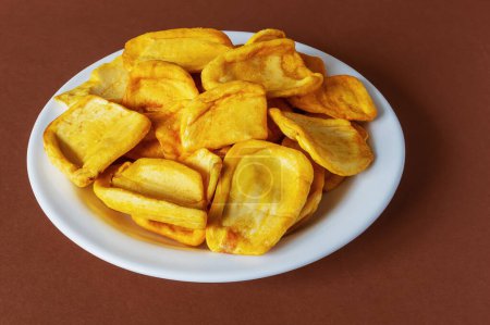 Keripik Sukun oder Breadfruit Chips ist ein Lebensmittel aus Brotfrüchten, das in dünne Scheiben geschnitten und gebraten wird, bis es trocken und knusprig ist. Serviert auf einem weißen Teller auf braunem Hintergrund