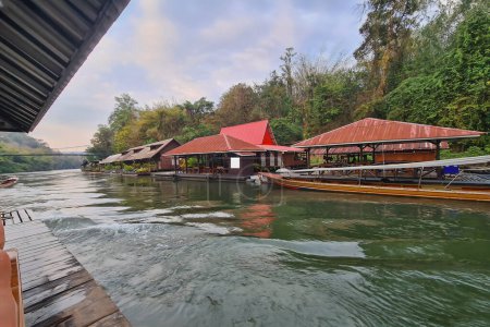 A tranquil riverside view of a peaceful resort with traditional red-roofed wooden houses on stilts over the water, the river reflecting the colors of the sky and small boats moored alongside pontoons.