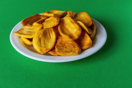Téléchargez les photos : Keripik Sukun ou chips de fruits à pain est un aliment à base de fruits à pain qui est finement tranché et frit jusqu'à ce que sec et croustillant. Servi sur une assiette blanche sur fond vert - en image libre de droit