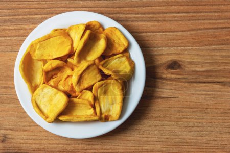 Téléchargez les photos : Keripik Sukun ou chips de fruits à pain est un aliment à base de fruits à pain qui est finement tranché et frit jusqu'à ce que sec et croustillant. Servi sur une assiette blanche sur une surface de table en pin - en image libre de droit
