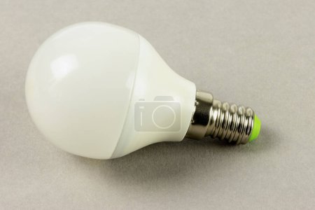 Die weiße LED-Lampe steht auf einem neutralen grauen Hintergrund, ihre undurchsichtige Kappe und der metallene Schraubfuß spiegeln moderne Energiespartechnologie wider..