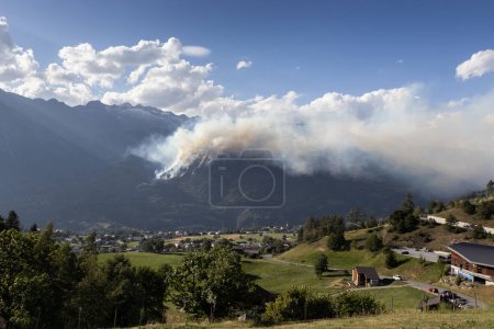 Foto de Los incendios forestales envuelven el lado de la montaña Riederhorn, cerca de Brig en Suiza con humo que llena el valle del Ródano. Incendio forestal real ocurrido en julio de 2023. - Imagen libre de derechos