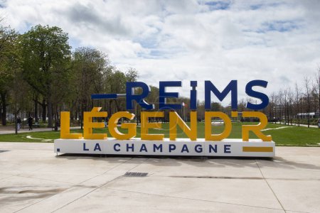 Foto de REIMS, FRANCIA, 3 DE ABRIL DE 2024: Vista de la calle de la ciudad de Reims y el parque de la ciudad con el letrero 'Reims' en letras azules grandes. Reims es un destino turístico popular en la zona de Champagne de Francia. - Imagen libre de derechos