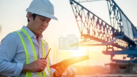 Téléchargez les photos : Homme ingénieur parlant sur talkie-walkie et utilisant une tablette pour travailler sur le chantier de construction au coucher du soleil - en image libre de droit