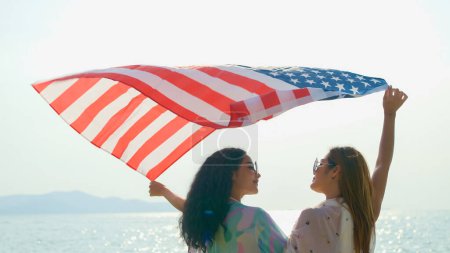 Foto de Mujeres jóvenes sostienen banderas estadounidenses en la playa y el mar en sus vacaciones de verano y sonríen y disfrutan de sus vacaciones. - Imagen libre de derechos