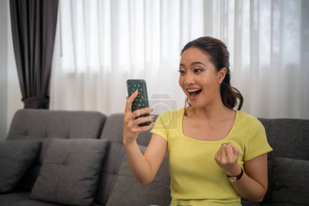 Téléchargez les photos : Asiatique femme assis sur canapé à la maison avec téléphone mobile et se sentir excité et heureux. - en image libre de droit
