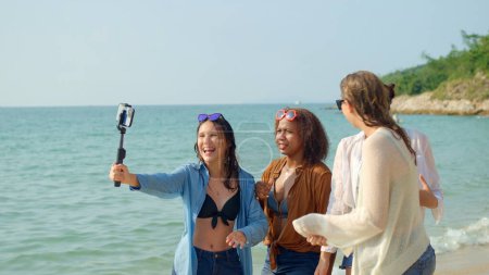 Foto de Un grupo de mujeres jóvenes usando teléfonos móviles para tomar selfies en la playa en sus vacaciones de verano y sonríen y disfrutan de sus vacaciones. - Imagen libre de derechos