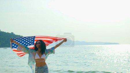 Foto de Mujeres jóvenes sostienen banderas estadounidenses en la playa y el mar en sus vacaciones de verano y sonríen y disfrutan de sus vacaciones. - Imagen libre de derechos