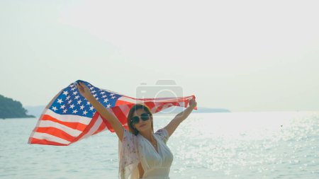 Téléchargez les photos : Jeunes femmes tiennent des drapeaux américains sur la plage et la mer pendant leurs vacances d'été et ils sourient et profitent de leurs vacances. - en image libre de droit