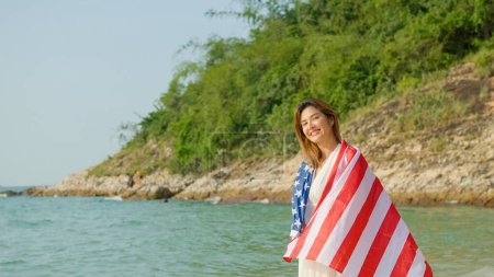 Téléchargez les photos : Jeunes femmes tiennent des drapeaux américains sur la plage et la mer pendant leurs vacances d'été et ils sourient et profitent de leurs vacances. - en image libre de droit