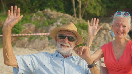 Téléchargez les photos : Un couple de personnes âgées assis sur des chaises à la plage regardant le soleil et la mer pendant leurs vacances d'été et ils sourient et profitent de leurs vacances. - en image libre de droit